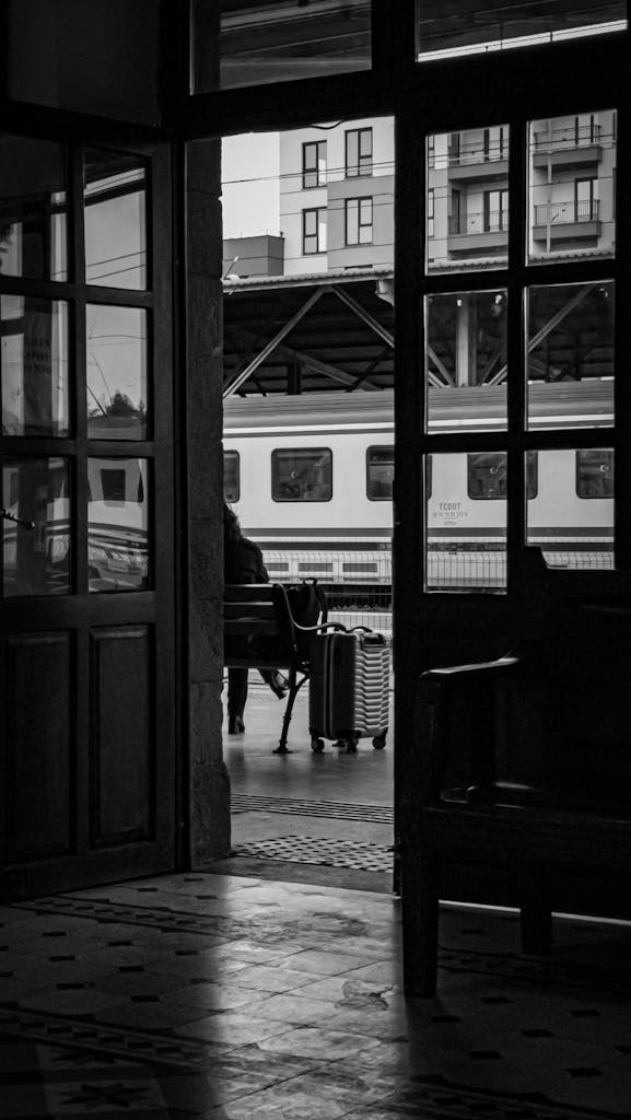 A person waits at a train station with a suitcase, framed by a doorway, in black and white.